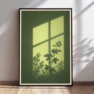 May include: A framed wall art print featuring a deep olive green background with a bright yellow rectangular light projection resembling a window pane. A dark silhouette of a leafy plant branch is centered within the light, casting shadows.