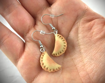 May include: A pair of silver dangle earrings with a light brown, crescent-shaped pastry charm. The pastry charm has a textured, crimped edge.