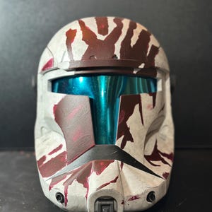 May include: A white and maroon helmet with a teal visor. The helmet has a weathered appearance with maroon paint splatters and streaks. The design is reminiscent of a military or science fiction aesthetic.