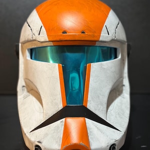 May include: A white and orange helmet with a blue visor. The helmet has black accents and a weathered appearance. The design features a prominent orange stripe across the top and front, with a black visor frame.