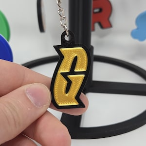 Puede incluir: Un llavero negro y dorado con un diseño estilizado de la letra "G". El llavero está sostenido en una mano, con otros llaveros coloridos y un soporte negro en el fondo. La "G" es dorada con un contorno negro.