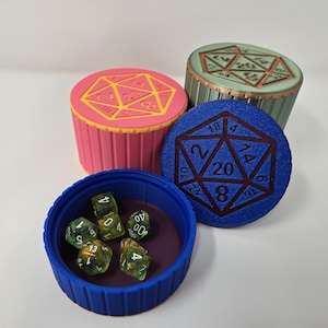 Puede incluir: Una colección de coloridos recipientes para dados y dados. Los recipientes son rosas, azules y verdes, cada uno con un diseño geométrico en la parte superior. El recipiente azul contiene varios dados translúcidos. Los dados son una mezcla de verde y amarillo.