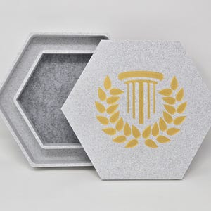 May include: A set of three hexagonal trays in shades of gray. The top tray features a gold-colored illustration of a column and laurel wreath. The trays are nested, with the largest tray on the bottom and the smallest on top.