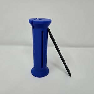 Puede incluir: Un objeto cilíndrico azul con una ranura y una herramienta rectangular negra apoyada contra él. El objeto tiene una parte superior e inferior redondeadas. La herramienta está en ángulo contra el lado del objeto azul.