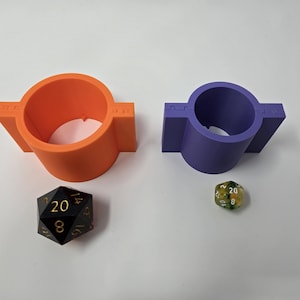 May include: Two dice holders, one orange and one purple, each with a circular opening and rectangular side extensions. A black and gold twenty-sided die and a green and yellow twenty-sided die are also present.