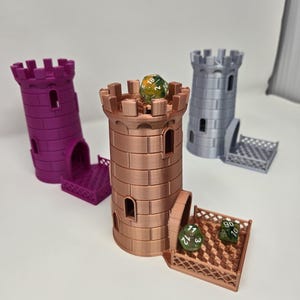 May include: Three 3D-printed dice towers in purple, copper, and gray. The copper tower has a dice on top and three dice in the tray. Each tower has a castle design with a crenelated top and arched windows. The towers are for tabletop gaming.