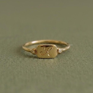 Op de afbeelding: Een goudkleurige ring met een rechthoekige plaat gegraveerd met de letter "K". De ring heeft twee kleine, ronde, heldere stenen aan weerszijden van de plaat. Op een groene achtergrond.