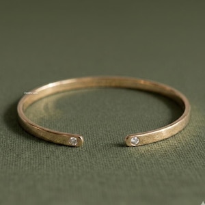 Könnte beinhalten: Ein goldfarbenes Armband in offener Form mit einem glatten, polierten Band, das an beiden abgerundeten Enden mit einem runden, klaren Edelstein besetzt ist. Das Metall weist eine feine, gebürstete Textur auf und hat eine breite Öffnung.