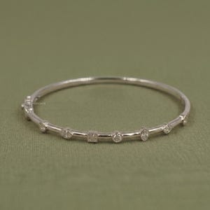 Op de afbeelding: Een zilveren armband met een reeks kleine, heldere edelstenen in verschillende vormen, waaronder rond, vierkant en marquise. De armband is geplaatst tegen een gedempte groene achtergrond.