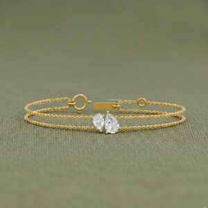 May include: A delicate gold bracelet featuring two clear, pear-shaped gemstones. The bracelet has a double chain design with a small clasp and a tag. The gemstones are the focal point, adding sparkle to the jewelry.
