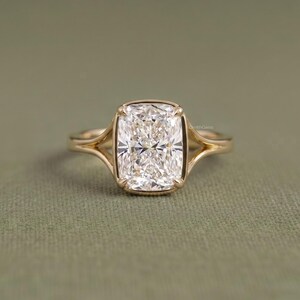 May include: A gold ring with a large, rectangular, clear gemstone. The gemstone is set in a bezel setting, and the band has a split shank design. The ring is displayed on a green surface. The text "DustinGems" is visible on the ring.