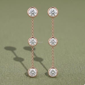 Op de afbeelding: Een paar roségouden oorbellen, elk voorzien van drie ronde, heldere, gefacetteerde stenen in een zetting. De stenen zijn verbonden door fijne roségouden kettinkjes, wat zorgt voor een verticaal, lineair ontwerp met een gepolijste metalen afwerking.