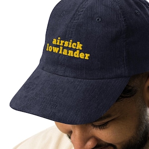 May include: Navy blue corduroy baseball cap with the text "airsick lowlander" embroidered in yellow. The cap has a curved brim and a structured fit. A stylish accessory for casual wear.