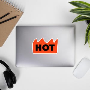May include: A sticker with the word "HOT" in bold black letters inside a red flame-shaped design. The sticker is on a laptop, with a notebook, pencils, headphones, a mouse, and a plant in the background.
