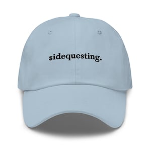 May include: A light blue baseball cap with the word "sidequesting" embroidered in black. The cap has a curved brim and an adjustable strap at the back. This hat is a casual accessory.