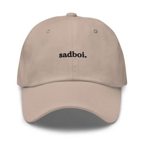 May include: A beige baseball cap with the word "sadboi." embroidered in black, centered on the front. The cap has a curved brim and a button on top. The hat is a neutral color, suitable for various outfits.