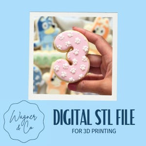 Op de afbeelding: Een roze koekje in de vorm van het cijfer drie, versierd met witte pootafdrukken. Het koekje wordt in een hand gehouden, met andere versierde koekjes op de achtergrond. De afbeelding bevat ook de tekst "DIGITAL STL FILE FOR 3D PRINTING".