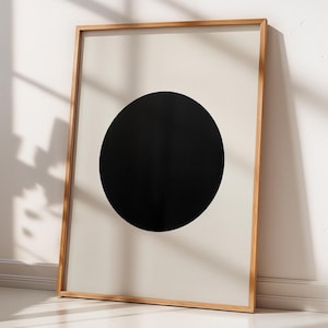 May include: A framed art print with a large black circle on a cream background. The artwork is in a light brown wooden frame, leaning against a white wall. A potted plant is nearby. The overall aesthetic is minimalist and modern.