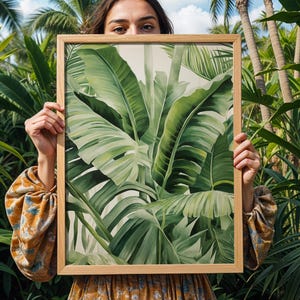 May include: A framed art print depicting large, green banana leaves. The artwork is presented in a natural wood frame, highlighting the tropical foliage. The print is held against a backdrop of dense green plants and palm trees.