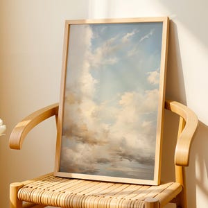 Könnte beinhalten: Ein gerahmtes Gemälde eines bewölkten Himmels in hellblauen und weißen Farbtönen. Das Gemälde hängt an einem Holzstuhl mit einem geflochtenen Sitz.
