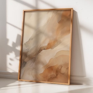 以下が含まれることがあります： 木製フレームに入った抽象的なアートプリント。アートワークは、ベージュ、茶色、タンの色が混ざり合い、水彩画のような効果を生み出しています。額装されたアートは、窓と鉢植えの植物の隣にある白い壁に立てかけられています。