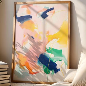 May include: Abstract art print in a wooden frame. The artwork features colourful brushstrokes in shades of pink, yellow, blue, and green. A stack of books and a potted plant are in the foreground, with a beige pillow and blanket.