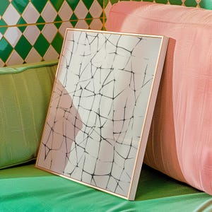Puede incluir: Impresión artística abstracta enmarcada, con una red de líneas negras sobre fondo blanco. El marco es dorado, apoyado contra un cojín rosa. El fondo incluye un cojín verde y una pared de azulejos verdes y blancos. Diseño moderno.