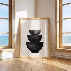 Puede incluir: Impresión artística enmarcada con un diseño minimalista de tres cuencos apilados de color gris oscuro sobre un fondo blanco. La obra de arte se exhibe en una habitación con luz natural y vista al océano.