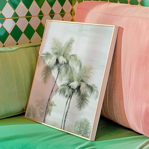 May include: A framed art print featuring a watercolor painting of palm trees. The artwork has a gold frame and a soft color palette of green, pink, and gray. The print is leaning against a pink cushion.