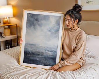 May include: An abstract landscape painting in a gold frame. The artwork depicts a cloudy sky and a body of water in shades of blue, gray, and white. The painting is held by a person sitting on a bed.