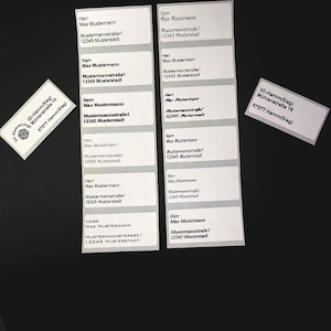 Könnte beinhalten: Eine Ansammlung weißer Adressetiketten mit schwarzer Schrift, angeordnet auf dunklem Untergrund. Die Etiketten tragen den Namen "Max Mustermann" und die Adresse "Mustermannstraße 1, 12345 Musterstadt". Ein kleines Etikett mit Logo und Adresse ist ebenfalls vorhanden.