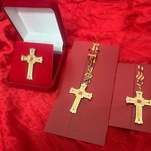 May include: Gold-coloured cross pendant with red gemstone accents, presented in a red velvet box and on red cards. The cross is detailed with a decorative design. The items are displayed on a red fabric background.
