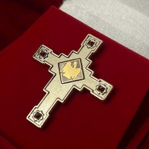 May include: A silver cross with a gold emblem at its centre, set with small red gemstones. The cross is presented on a red velvet surface, possibly a jewellery or religious item.