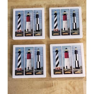 May include: A set of four square coasters, each with a lighthouse design. The coasters show three lighthouse illustrations: one with black and white stripes, one with a red top, and one with black stripes.
