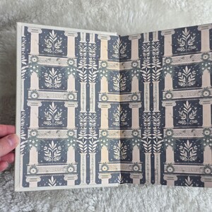 May include: Open book with a decorative pattern featuring white columns, floral elements, and a dark blue background. The design is symmetrical and detailed, with a classical aesthetic. The book's pages are open, revealing the interior design.