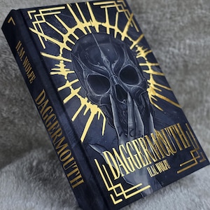 May include: A hardcover book titled Dagger-Mouth by H.M. Wolfe. The black cover features a central illustration of a dark skull with gold foil accents, including a radiating halo design and geometric borders. The title is printed in gold foil.