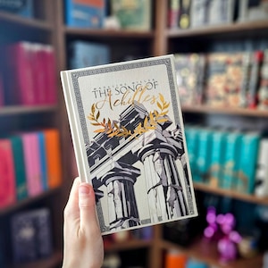 May include: A hardcover book titled "The Song of Achilles" by Madeline Miller. The cover features a black and white illustration of Greek columns with gold accents and a decorative border. The book is held in front of a bookshelf.