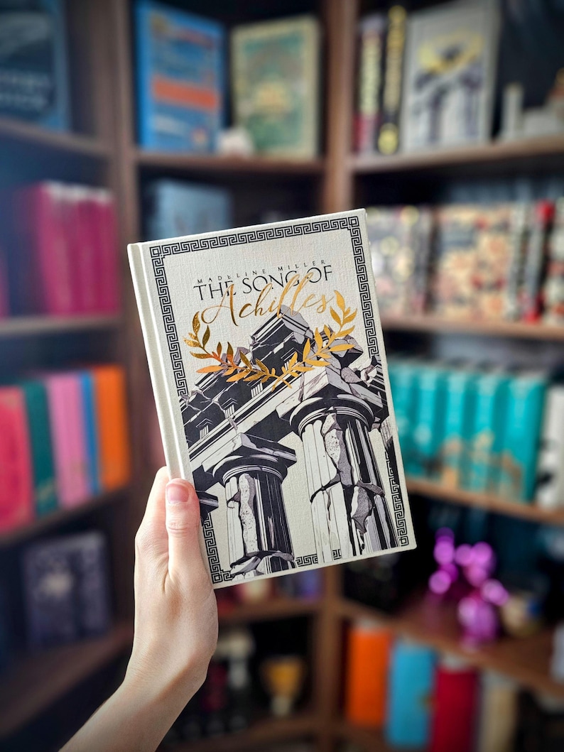 May include: A hardcover book titled "The Song of Achilles" by Madeline Miller. The cover features a black and white illustration of Greek columns with gold accents and a decorative border. The book is held in front of a bookshelf.