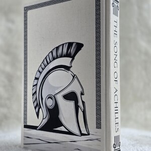 May include: A hardcover book titled "The Song of Achilles" with a black and white illustration of a Spartan helmet on the cover. The cover has a decorative border and the spine displays the title vertically.