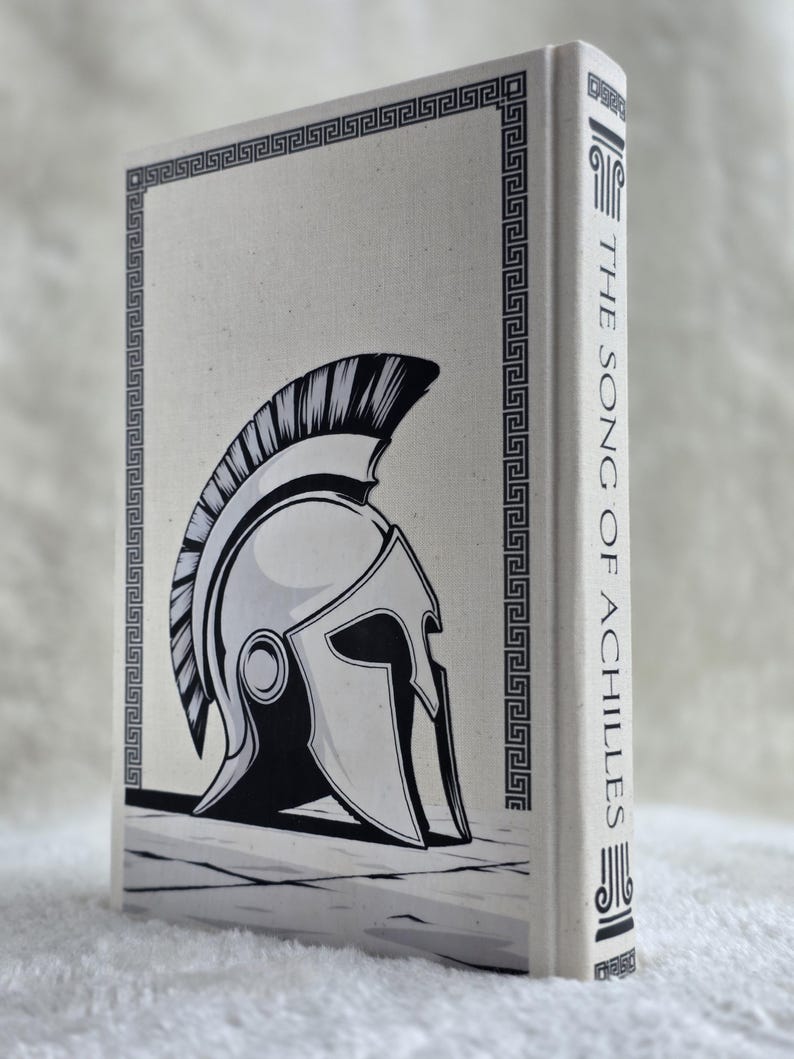 May include: A hardcover book titled "The Song of Achilles" with a black and white illustration of a Spartan helmet on the cover. The cover has a decorative border and the spine displays the title vertically.