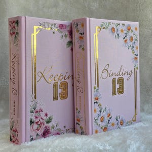 May include: Two pink hardcover books with gold lettering and floral designs. One book reads "Keeping 13" and the other reads "Binding 13". The books have gold frames and are decorated with flowers.