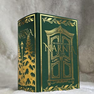 May include: A hardcover book titled "The Chronicles of Narnia" by C.S. Lewis. The book has a dark green cover with gold foil lettering and illustrations, including a wardrobe and a winter scene with a lamppost and trees. The book is displayed on a soft, white surface.
