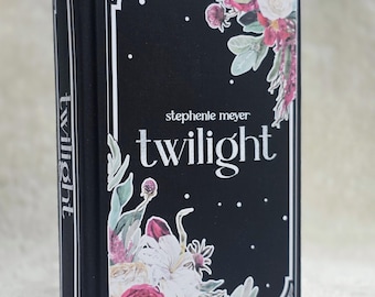 May include: A hardcover book titled "Twilight" by Stephenie Meyer. The book has a black cover with white text and floral illustrations in shades of pink, white, and green. The spine also features the word "Twilight" in white.
