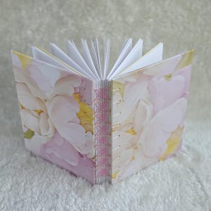 May include: A handmade journal with a floral design. The cover features a pattern of pink and white flowers with yellow accents. The pages are white, and the spine is stitched with pink thread.
