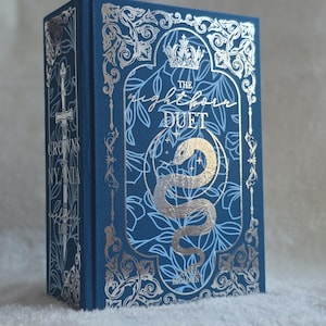 May include: A navy blue hardcover book with silver foil detailing. The cover reads "The Nightborn Duet" with a snake and crown design. The spine shows "Crowns Nyaxia Duet". The book has decorative floral patterns.