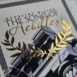 May include: A book cover featuring the title "The Song of Achilles" in gold script. The cover has a black and white illustration of classical architecture and a gold laurel wreath. The author's name, Madeline Miller, is also visible.