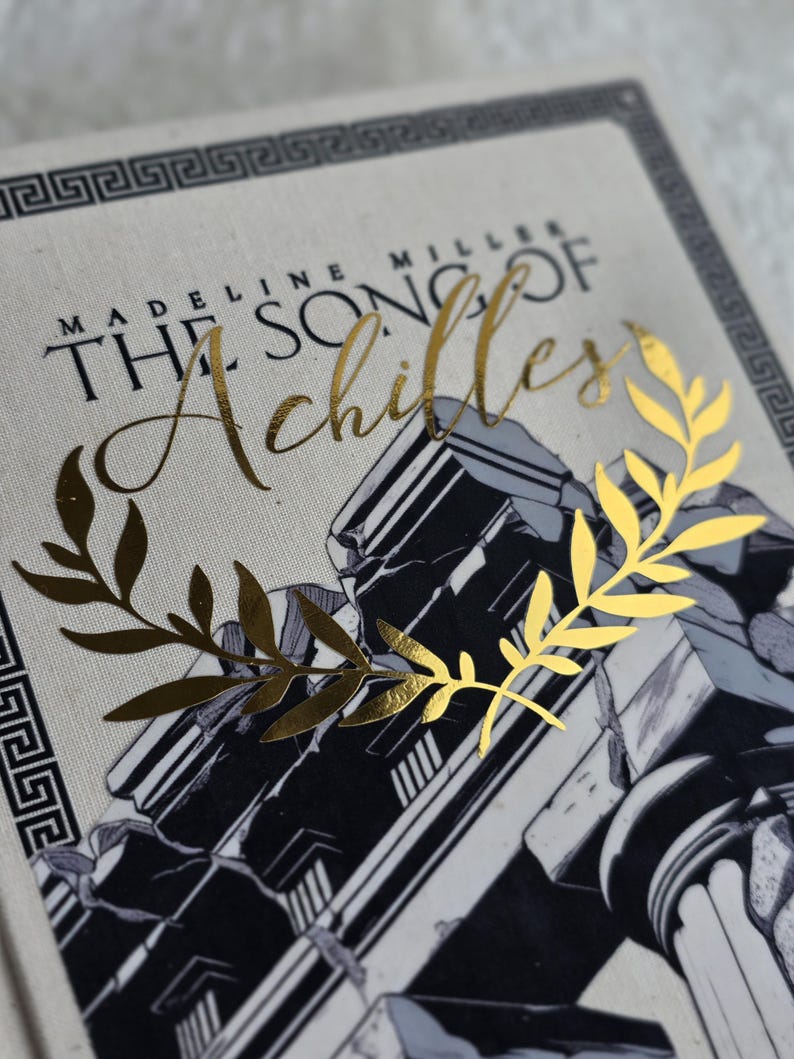 May include: A book cover featuring the title "The Song of Achilles" in gold script. The cover has a black and white illustration of classical architecture and a gold laurel wreath. The author's name, Madeline Miller, is also visible.