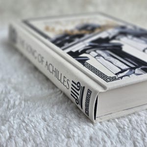 May include: A hardcover book titled "The Song of Achilles" with a white cover and spine. The cover features a black and white illustration of classical architecture with gold accents. The spine displays the title in black text.