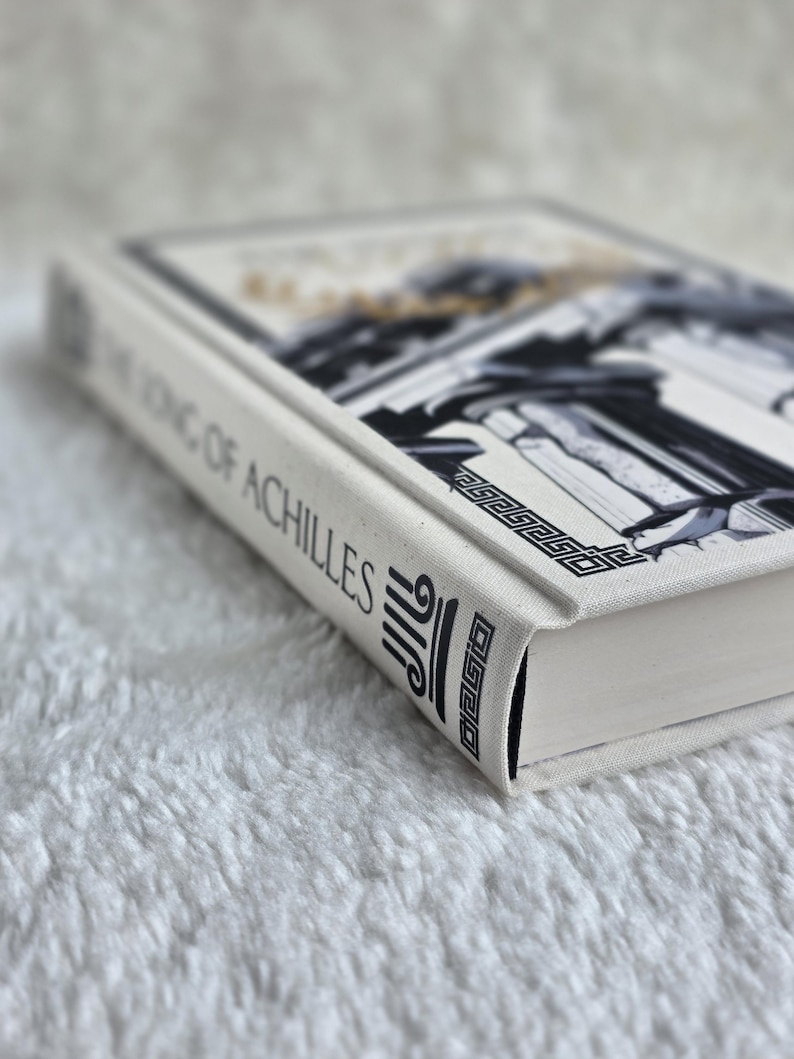May include: A hardcover book titled "The Song of Achilles" with a white cover and spine. The cover features a black and white illustration of classical architecture with gold accents. The spine displays the title in black text.