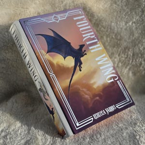 Könnte beinhalten: Ein gebundenes Buch mit dem Titel Fourth Wing von Rebecca Yarros. Das Cover zeigt die Silhouette eines dunklen Drachen vor einem Sonnenuntergangshimmel mit Wolken, umrahmt von silbernen geometrischen Linien. Der Buchrücken zeigt den Titel.